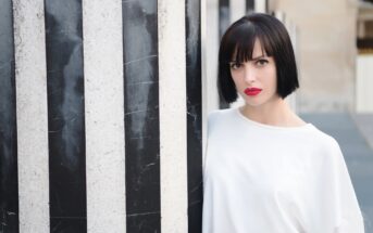 A woman with short black hair and red lipstick stands behind a large black-and-white striped column, gazing directly at the camera. She is wearing a white top and the background appears to be an outdoor urban setting.