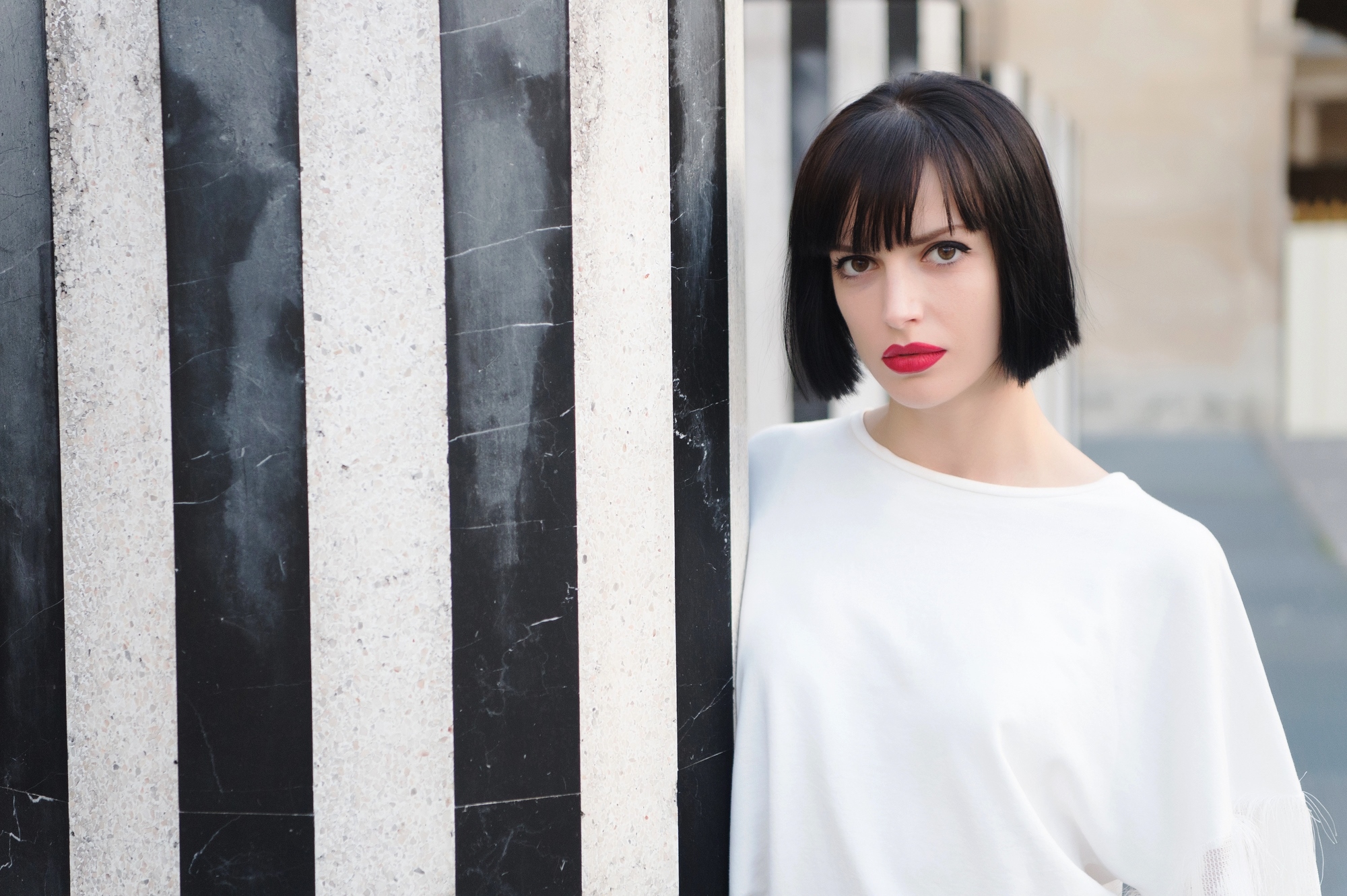 A woman with short black hair and red lipstick stands behind a large black-and-white striped column, gazing directly at the camera. She is wearing a white top and the background appears to be an outdoor urban setting.