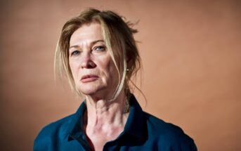 A woman with light-colored hair styled loosely and wearing a blue shirt gazes ahead with a serious expression. The background is a solid, muted brown color.