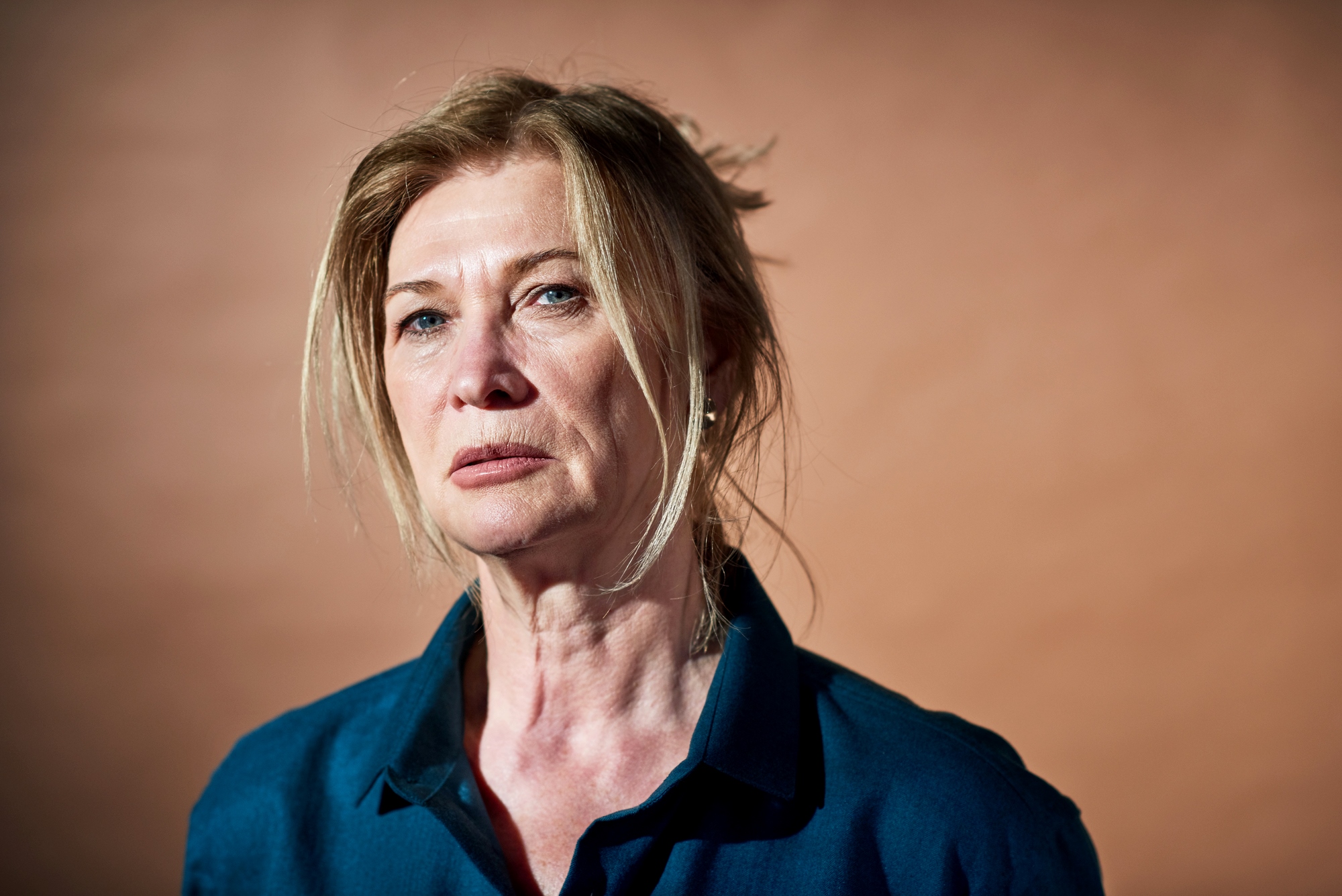 A woman with light-colored hair styled loosely and wearing a blue shirt gazes ahead with a serious expression. The background is a solid, muted brown color.