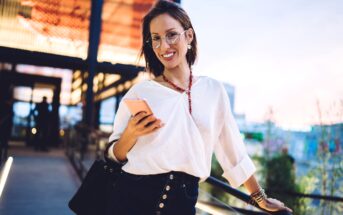 A woman with glasses and short brown hair is smiling and holding a smartphone. She is wearing a white blouse and a black skirt, standing outdoors with a modern building in the background, as well as a slightly blurred person in the distance.