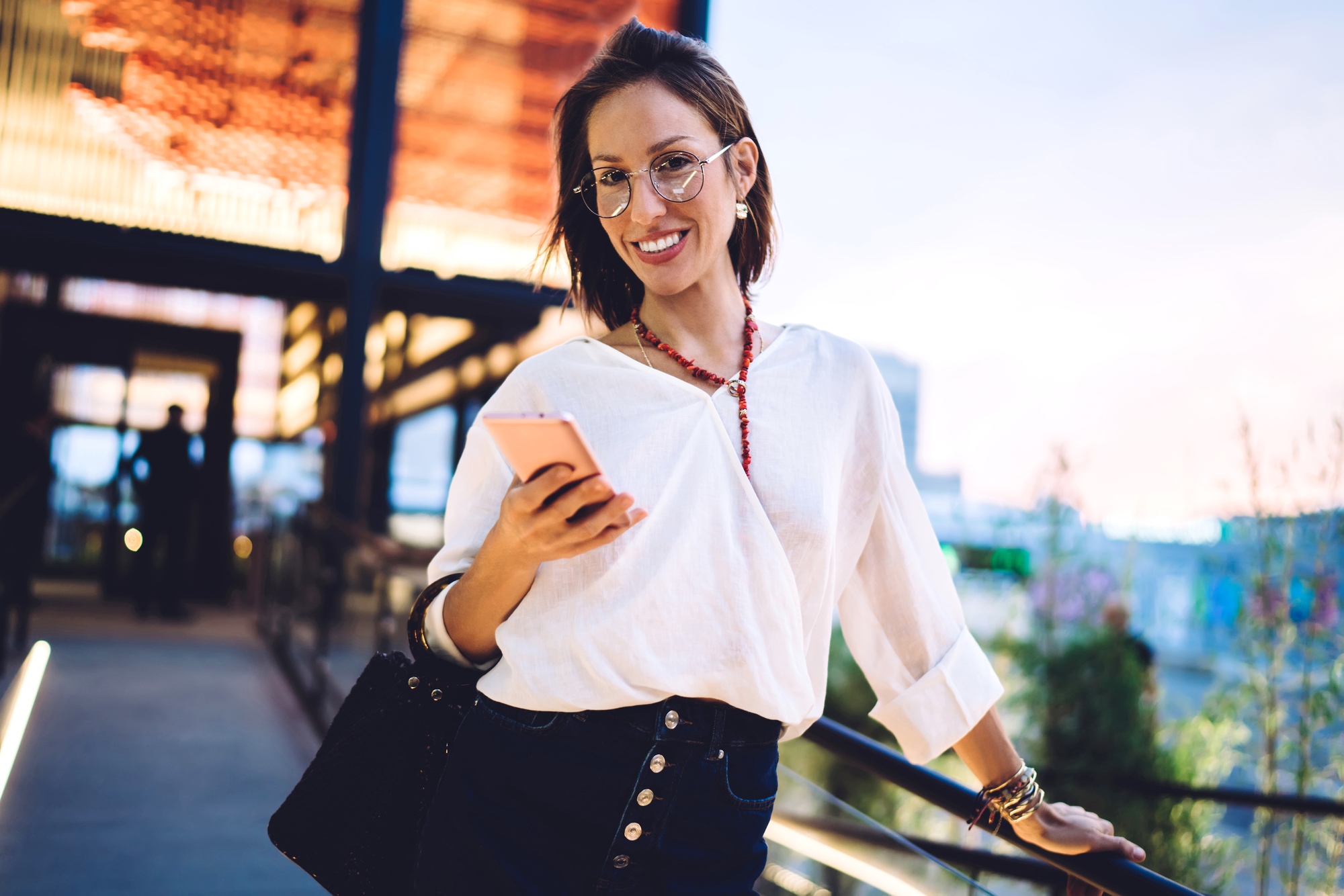 A woman with glasses and short brown hair is smiling and holding a smartphone. She is wearing a white blouse and a black skirt, standing outdoors with a modern building in the background, as well as a slightly blurred person in the distance.