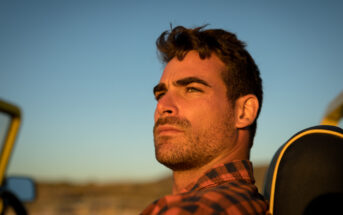 A man with a serious expression sits in a vehicle with the top down, bathed in warm sunlight. He wears a plaid shirt and has short, tousled hair and a beard. The background features an out-of-focus landscape with a clear sky at sunset.