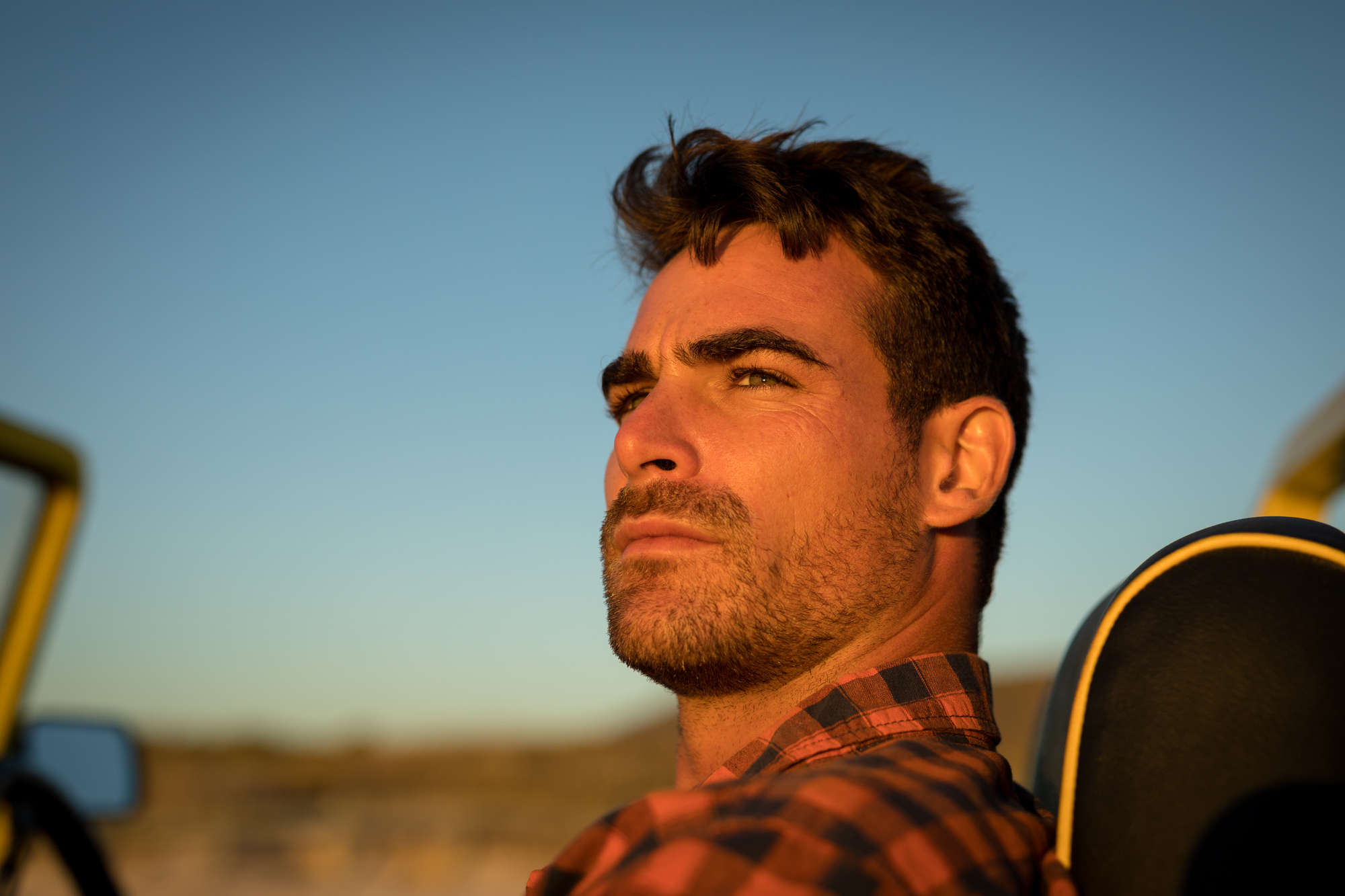 A man with a serious expression sits in a vehicle with the top down, bathed in warm sunlight. He wears a plaid shirt and has short, tousled hair and a beard. The background features an out-of-focus landscape with a clear sky at sunset.