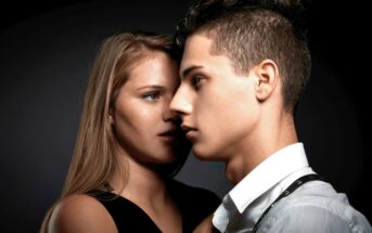 Two individuals stand close to each other against a dark background. The person on the left has long blonde hair and is whispering into the ear of the person on the right, who has short hair styled upwards and is looking ahead with a serious expression.