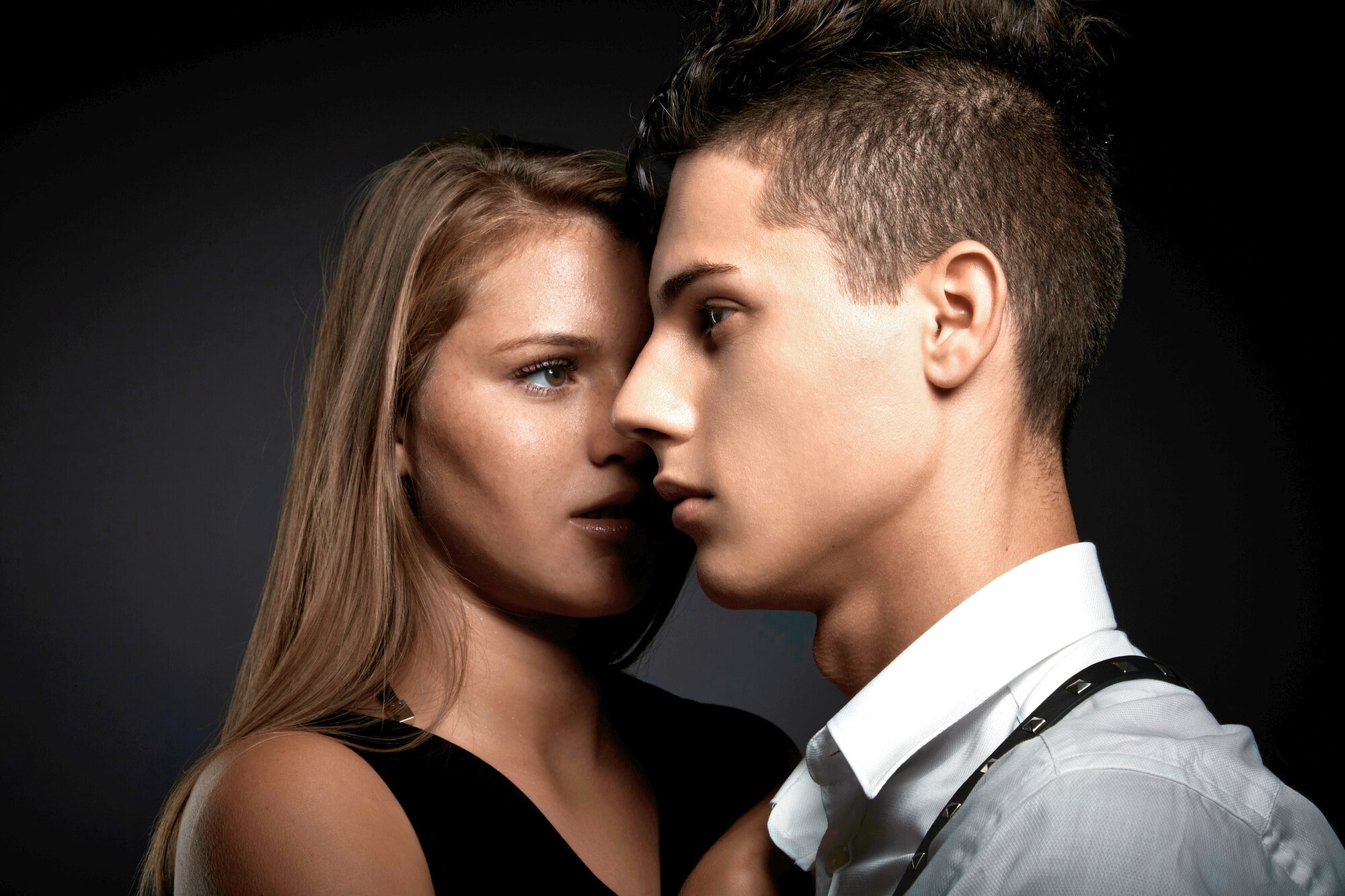 Two individuals stand close to each other against a dark background. The person on the left has long blonde hair and is whispering into the ear of the person on the right, who has short hair styled upwards and is looking ahead with a serious expression.