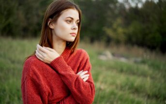 A young woman with long brown hair stands in a grassy field. She is wearing a red sweater and looking off to the side with a thoughtful expression, her arms crossed and one hand touching her opposite shoulder. The background features blurred greenery.