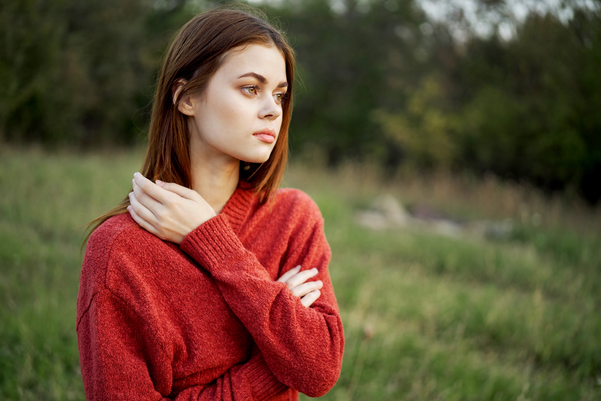 A young woman with long brown hair stands in a grassy field. She is wearing a red sweater and looking off to the side with a thoughtful expression, her arms crossed and one hand touching her opposite shoulder. The background features blurred greenery.