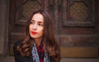 Woman with long, wavy brown hair and red lipstick, wearing a black coat over a red plaid shirt and a paisley scarf, looking slightly to the side. She is standing in front of a wooden door with ornate hexagonal patterns.