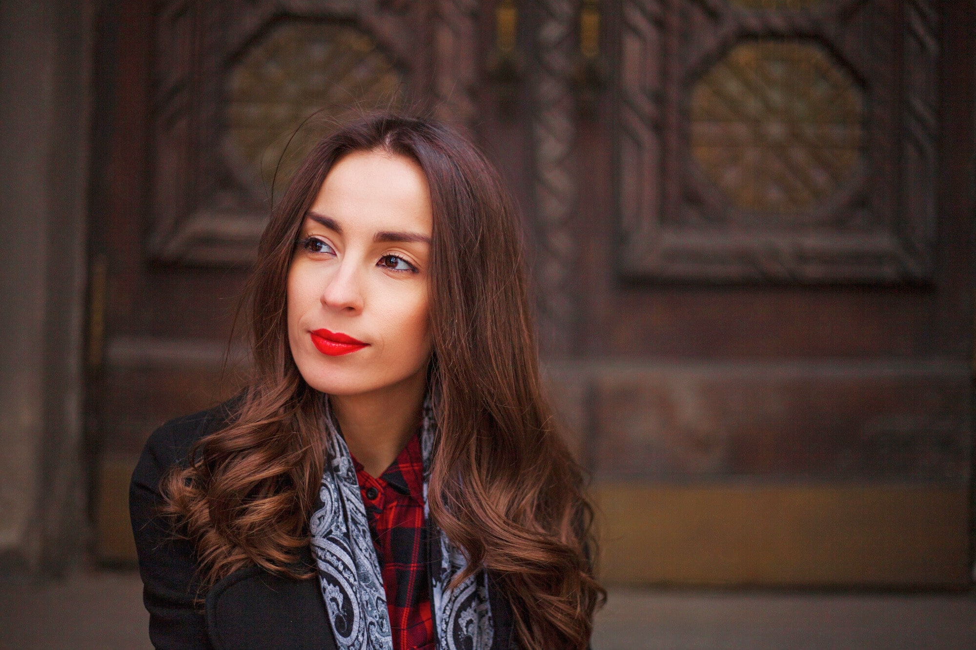 Woman with long, wavy brown hair and red lipstick, wearing a black coat over a red plaid shirt and a paisley scarf, looking slightly to the side. She is standing in front of a wooden door with ornate hexagonal patterns.