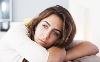 A young woman with long brown hair and blue eyes rests her head on her folded arms, gazing pensively into the distance. She wears a white long-sleeved top and appears to be deep in thought. The background is softly blurred.