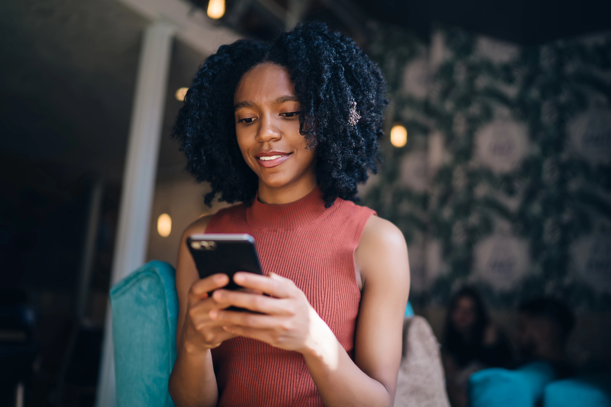 A woman with curly hair, wearing a sleeveless orange top, is smiling while looking at her smartphone. She is seated in a cozy, dimly lit room with patterned wallpaper and soft furniture.