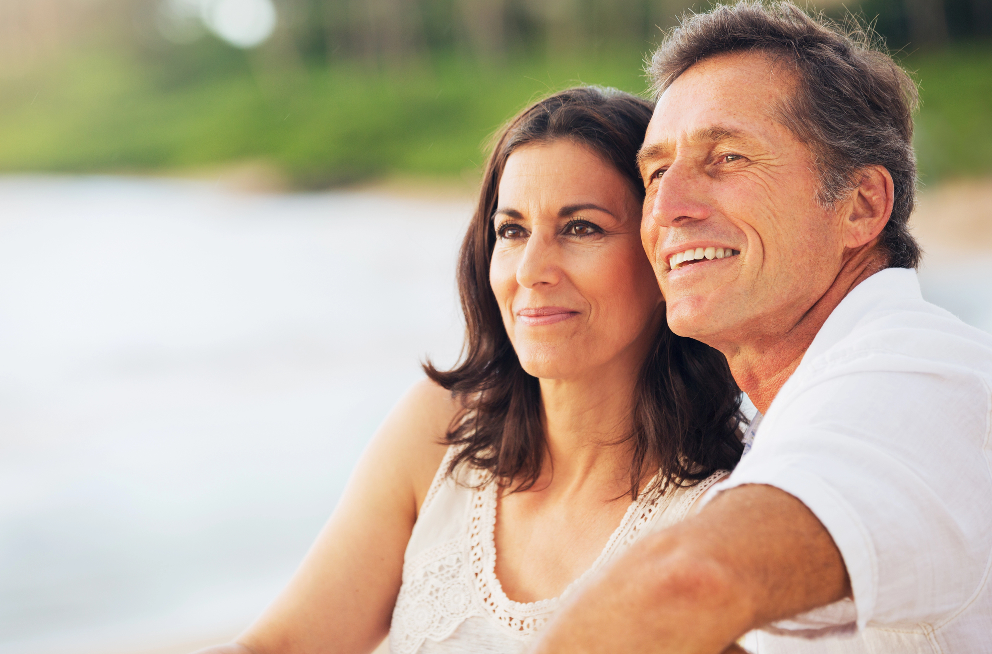 A man and woman sit close together outdoors, both smiling and gazing into the distance. The woman has long brown hair and is wearing a white top, while the man has short gray hair and is dressed in a white shirt. Blurred greenery is visible in the background.