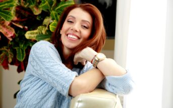 A woman with long, auburn hair smiles brightly while resting her arms on a beige couch. She wears a light blue sweater and a gold watch. In the background, there is a leafy green plant with red accents illuminated by natural light from a nearby window.