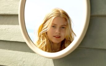 A young woman with long, blonde hair is reflected in a round mirror. She appears to be outdoors, standing in front of light-green wooden planks. She has a relaxed expression on her face, and the sky is lightly visible in the background.