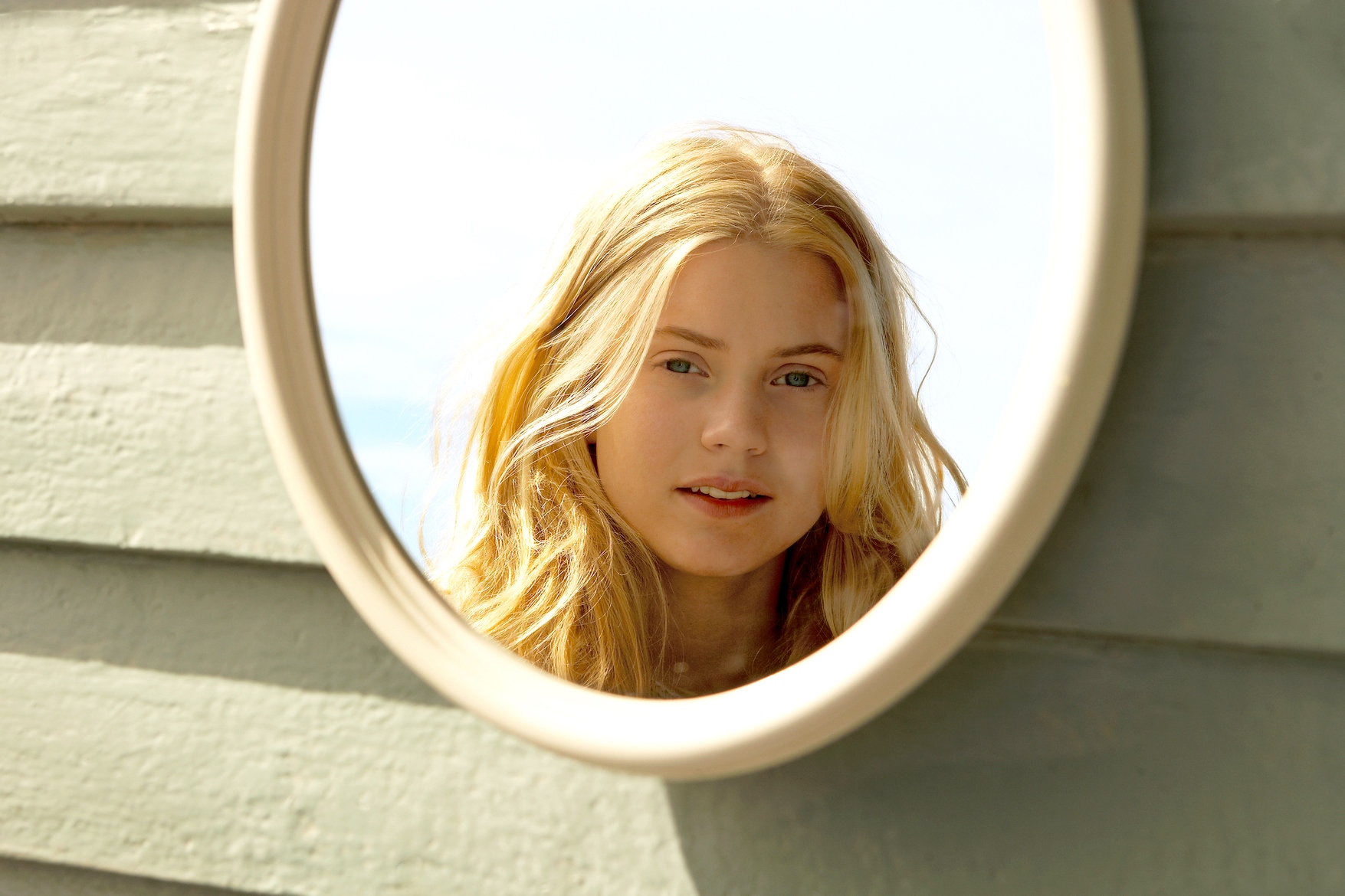 A young woman with long, blonde hair is reflected in a round mirror. She appears to be outdoors, standing in front of light-green wooden planks. She has a relaxed expression on her face, and the sky is lightly visible in the background.