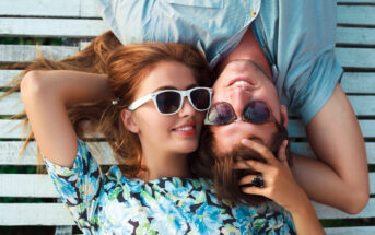 A woman and a man lay side by side on a wooden deck, both wearing sunglasses. The woman wears a floral top, and the man is in a blue shirt. They are smiling and look relaxed. The woman is resting her hand on the man's head.