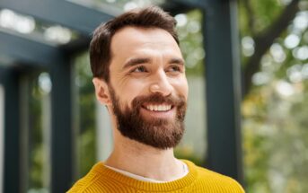 A smiling man with a full beard and mustache is gazing slightly upwards. He is wearing a yellow sweater and has short, neatly styled hair. The background is blurred with green foliage and a structure suggesting an outdoor setting.