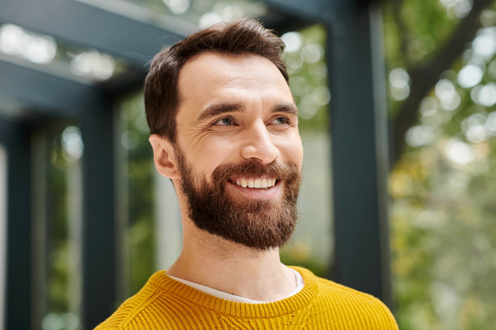 A smiling man with a full beard and mustache is gazing slightly upwards. He is wearing a yellow sweater and has short, neatly styled hair. The background is blurred with green foliage and a structure suggesting an outdoor setting.