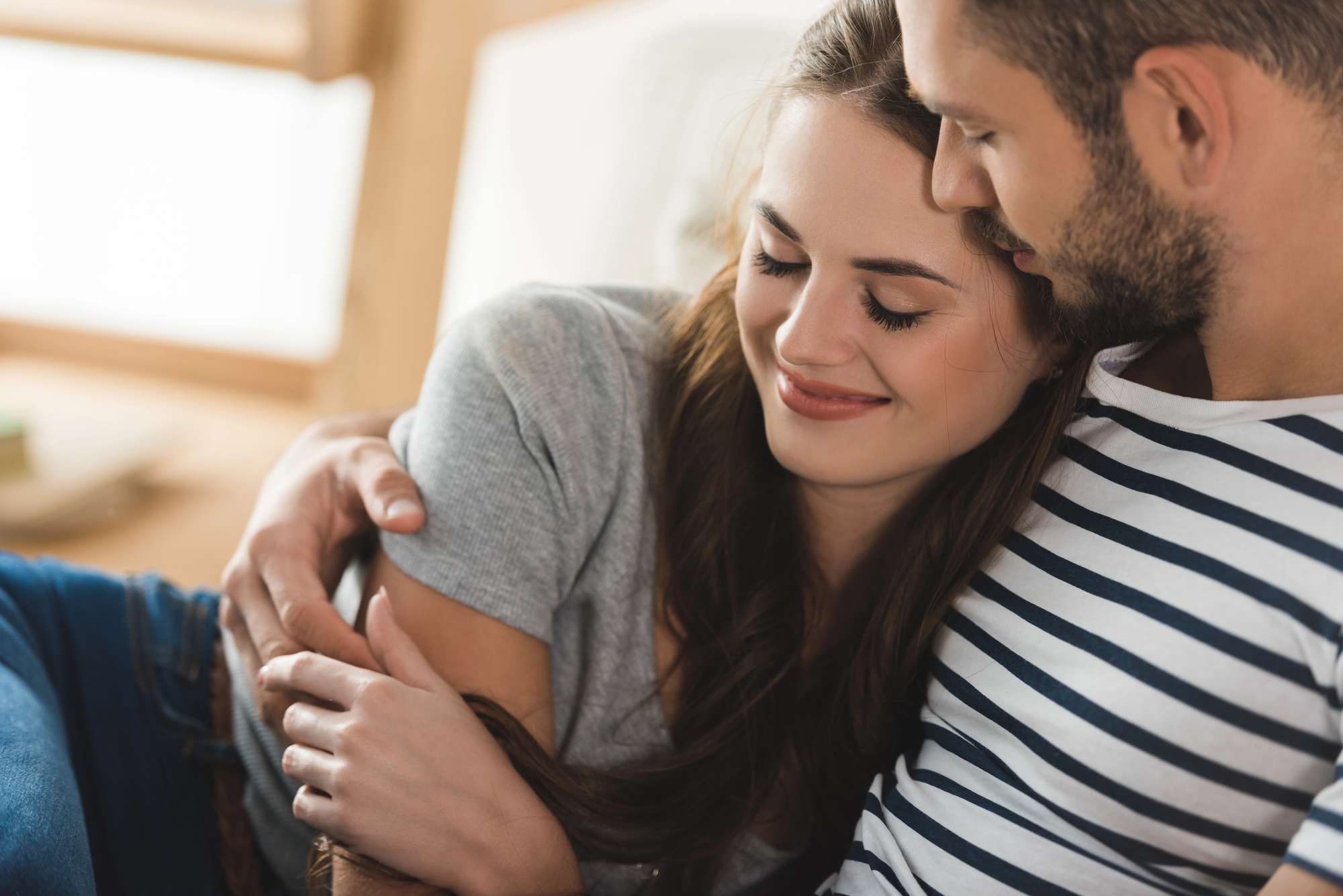 A young couple is sitting closely together on a sofa. The woman has long brown hair and is wearing a gray shirt, leaning her head on the man's shoulder with a contented smile. The man is wearing a striped shirt and has his arm wrapped around her, looking down at her affectionately.