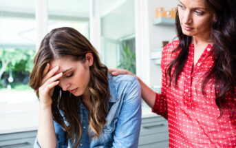 A woman in a denim shirt holds her forehead in distress while another woman in a red blouse stands beside her, offering comfort with a hand on her shoulder. They are in a bright room with natural light coming through a window in the background.