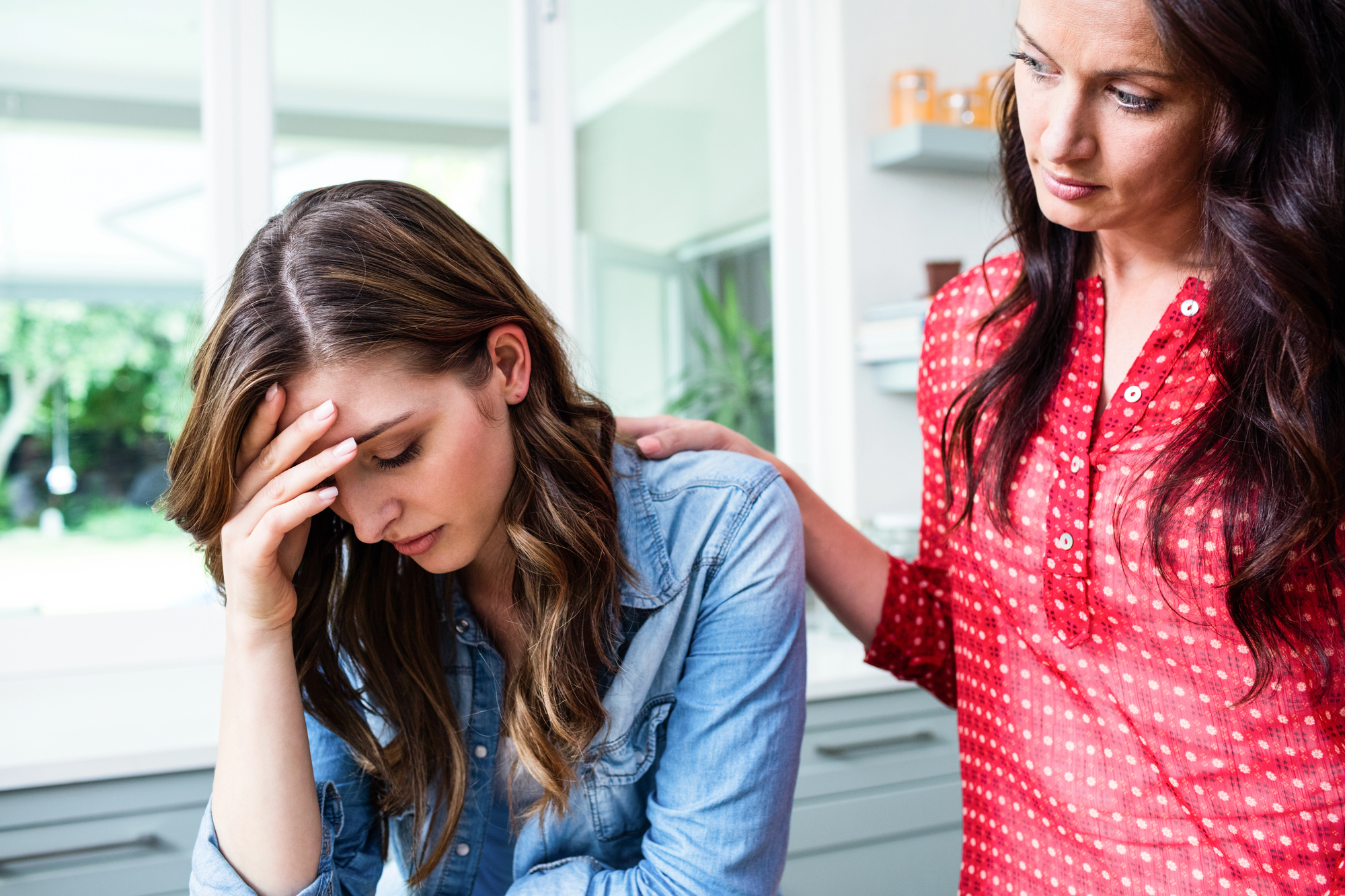 A woman in a denim shirt holds her forehead in distress while another woman in a red blouse stands beside her, offering comfort with a hand on her shoulder. They are in a bright room with natural light coming through a window in the background.