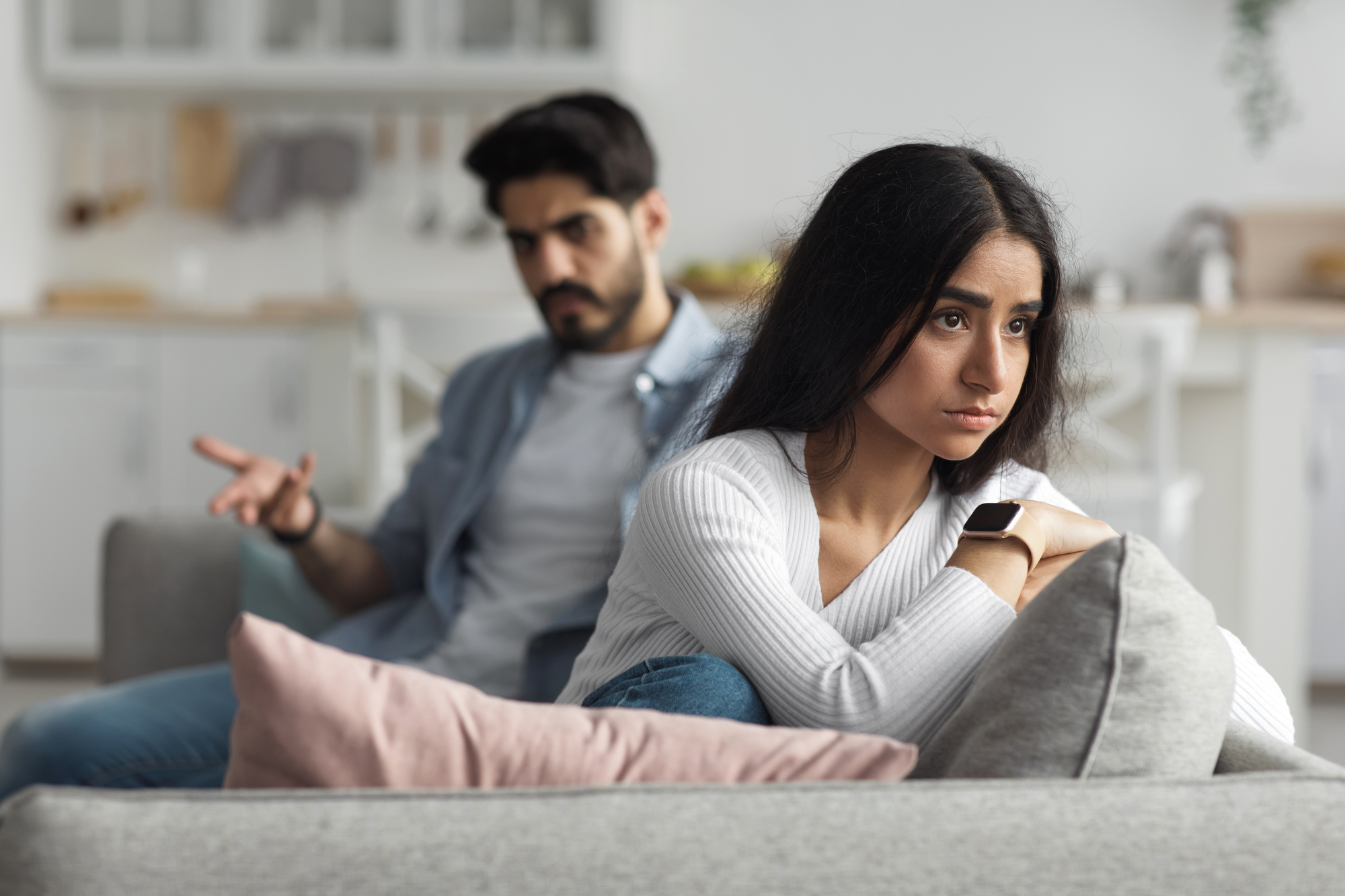 A young woman sits on a couch with a distressed expression, arms folded, staring into the distance. Behind her, a man with a frustrated look gestures with one hand, seemingly trying to communicate. The background shows a modern kitchen.