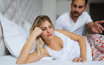 A woman with blonde hair lies on her side in bed, looking down and appearing upset. A man with short dark hair sits beside her, gesturing with his hand as if trying to communicate. Both are dressed in casual white clothing, and the bed is white.