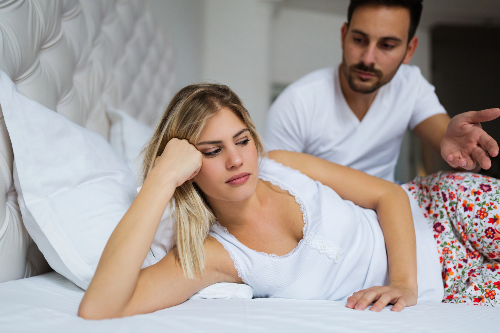 A woman with blonde hair lies on her side in bed, looking down and appearing upset. A man with short dark hair sits beside her, gesturing with his hand as if trying to communicate. Both are dressed in casual white clothing, and the bed is white.