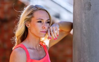 A woman with long blonde hair, wearing a bright pink sleeveless top, is leaning against a gray pillar. She has her right arm bent and resting on the pillar, with her hand near her face. She gazes thoughtfully into the distance.