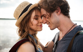 A young couple enjoys a tender moment on a beach. The woman, wearing a straw hat, gently touches foreheads with the man, who is smiling warmly. They both have their eyes closed and appear to be content. The beach and water are blurred out in the background.