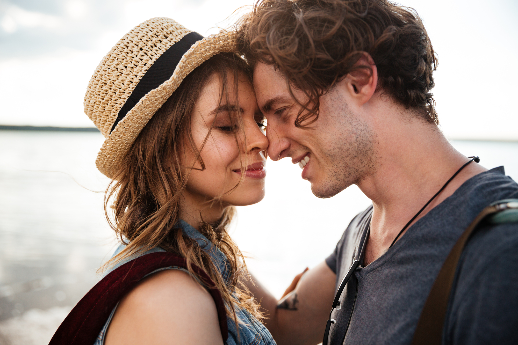 A young couple enjoys a tender moment on a beach. The woman, wearing a straw hat, gently touches foreheads with the man, who is smiling warmly. They both have their eyes closed and appear to be content. The beach and water are blurred out in the background.