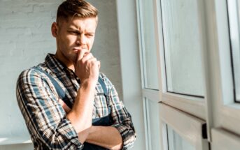 A man with short brown hair, wearing a checkered shirt and a dark apron, stands thoughtfully by a window with his hand on his chin. He appears to be deep in thought, gazing outside as sunlight streams in through the window.