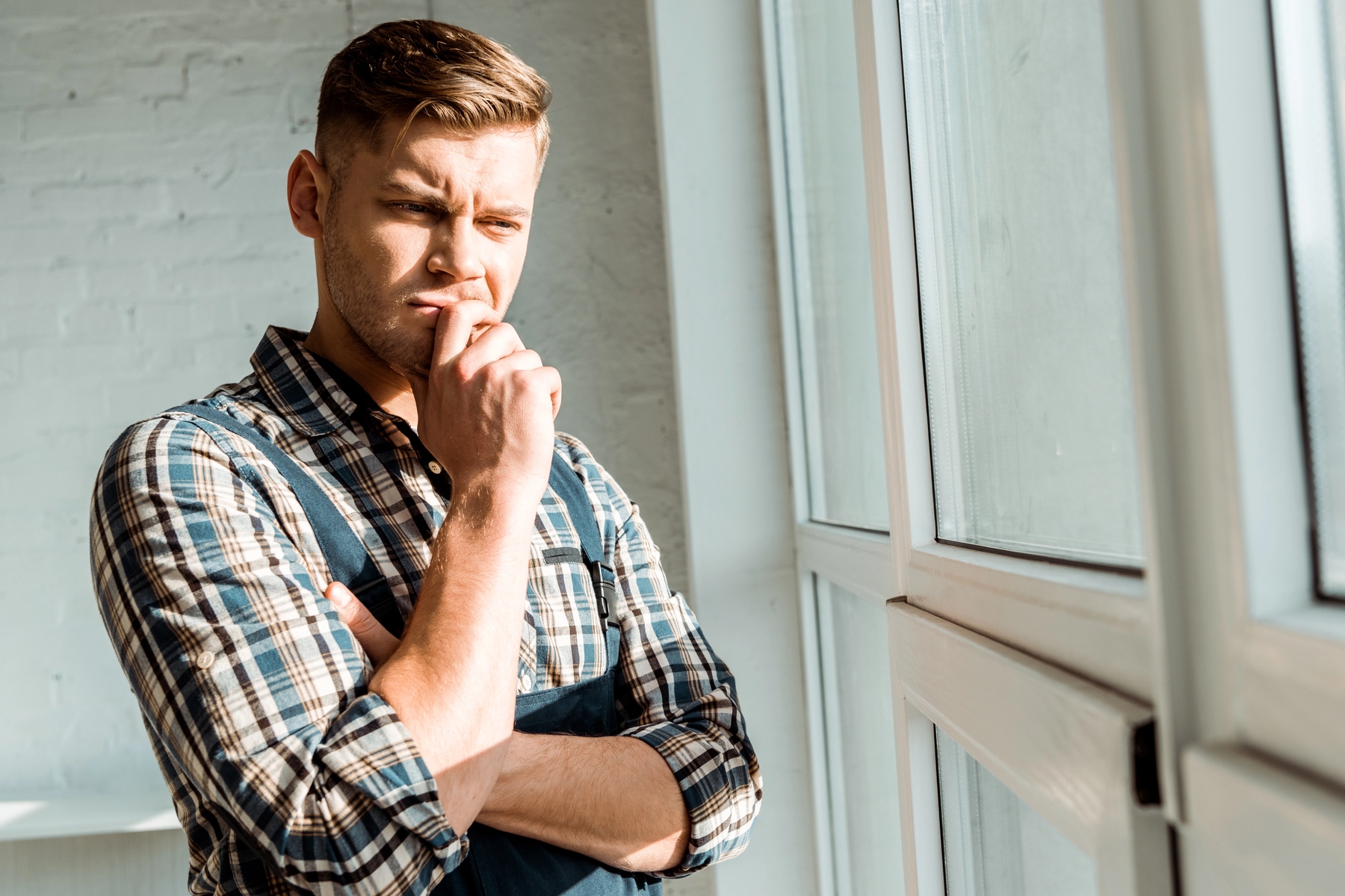 A man with short brown hair, wearing a checkered shirt and a dark apron, stands thoughtfully by a window with his hand on his chin. He appears to be deep in thought, gazing outside as sunlight streams in through the window.