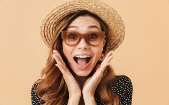 A woman with long wavy hair wearing a straw hat and brown-framed sunglasses, has her hands on her cheeks and an excited expression. She is dressed in a black polka-dot top and is set against a solid peach-colored background.