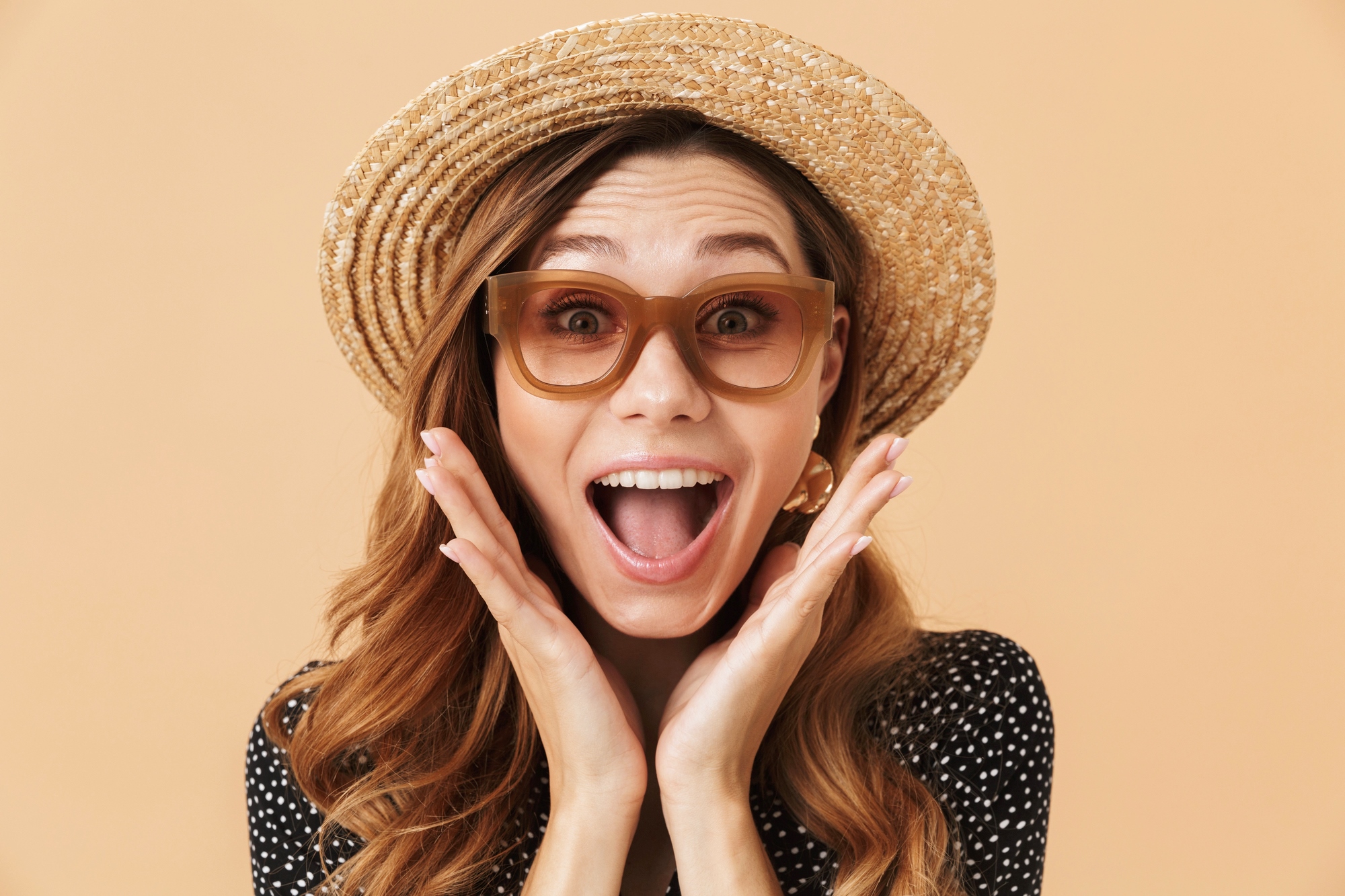 A woman with long wavy hair wearing a straw hat and brown-framed sunglasses, has her hands on her cheeks and an excited expression. She is dressed in a black polka-dot top and is set against a solid peach-colored background.