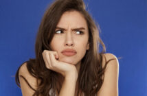 A woman with long brown hair is frowning and resting her chin on her hand while looking off to the side. She has a puzzled and slightly annoyed expression on her face. The background is a solid blue color.