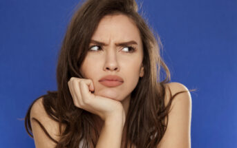 A woman with long brown hair is frowning and resting her chin on her hand while looking off to the side. She has a puzzled and slightly annoyed expression on her face. The background is a solid blue color.