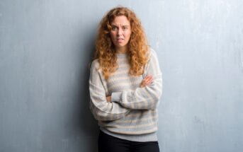 A woman with curly red hair stands against a light gray textured wall. She is wearing a light gray and white striped sweater and black pants. Her arms are crossed, and she has a somewhat confused or uncertain expression on her face.