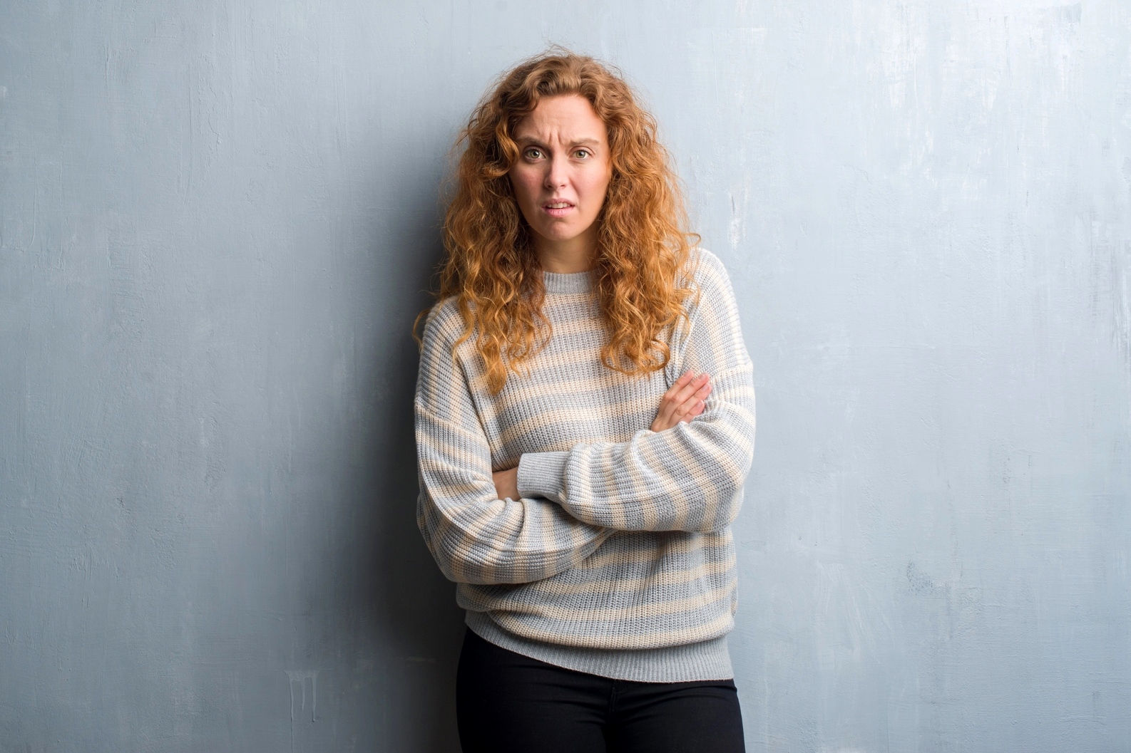 A woman with curly red hair stands against a light gray textured wall. She is wearing a light gray and white striped sweater and black pants. Her arms are crossed, and she has a somewhat confused or uncertain expression on her face.