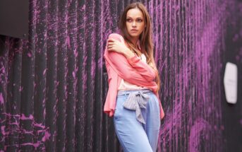 A woman with long hair stands against a black wall with pink abstract streaks. She wears a light pink blouse and blue pants tied with a bow in front. She has one arm crossed over her chest and looks off to the side with a thoughtful expression.