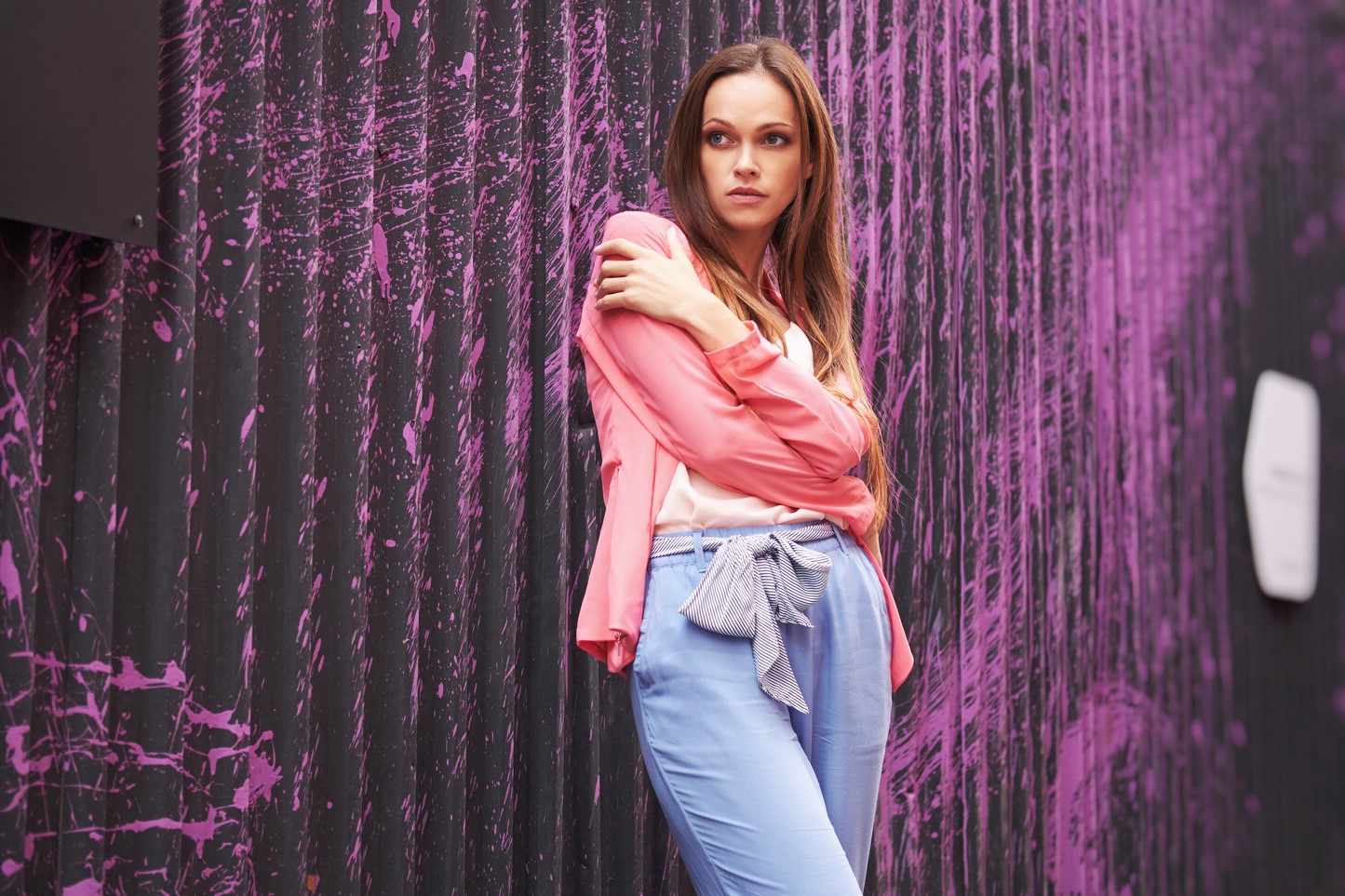 A woman with long hair stands against a black wall with pink abstract streaks. She wears a light pink blouse and blue pants tied with a bow in front. She has one arm crossed over her chest and looks off to the side with a thoughtful expression.