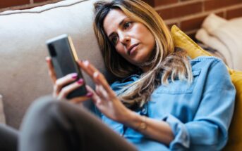 A woman in a denim shirt lies on a couch, looking at her smartphone. She appears thoughtful or pensive, with a relaxed posture. A brick wall is visible in the background.