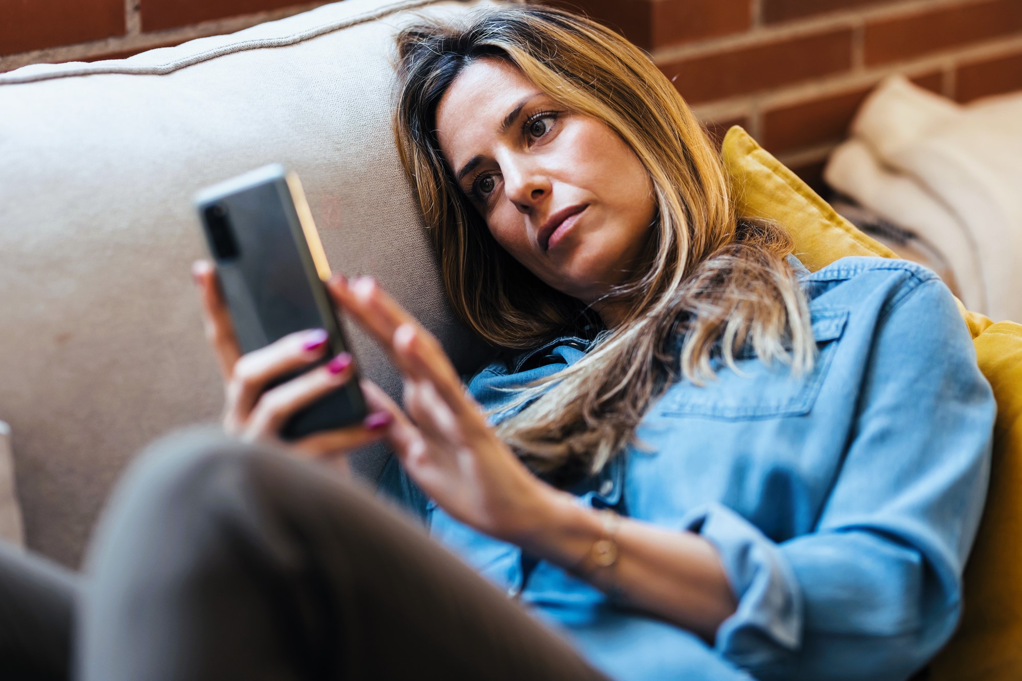 A woman in a denim shirt lies on a couch, looking at her smartphone. She appears thoughtful or pensive, with a relaxed posture. A brick wall is visible in the background.