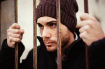 A person with a knitted brown beanie and a nose piercing looks through rusty iron bars, gripping them with both hands. The individual has a focused expression and is clad in dark clothing. The background is blurred.