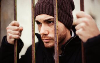 A person with a knitted brown beanie and a nose piercing looks through rusty iron bars, gripping them with both hands. The individual has a focused expression and is clad in dark clothing. The background is blurred.