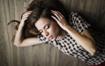 A woman with long brown hair is lying on the wooden floor with her eyes closed. She is wearing a patterned top with circular designs and has her hands gently placed on the sides of her head. The background is also wooden.