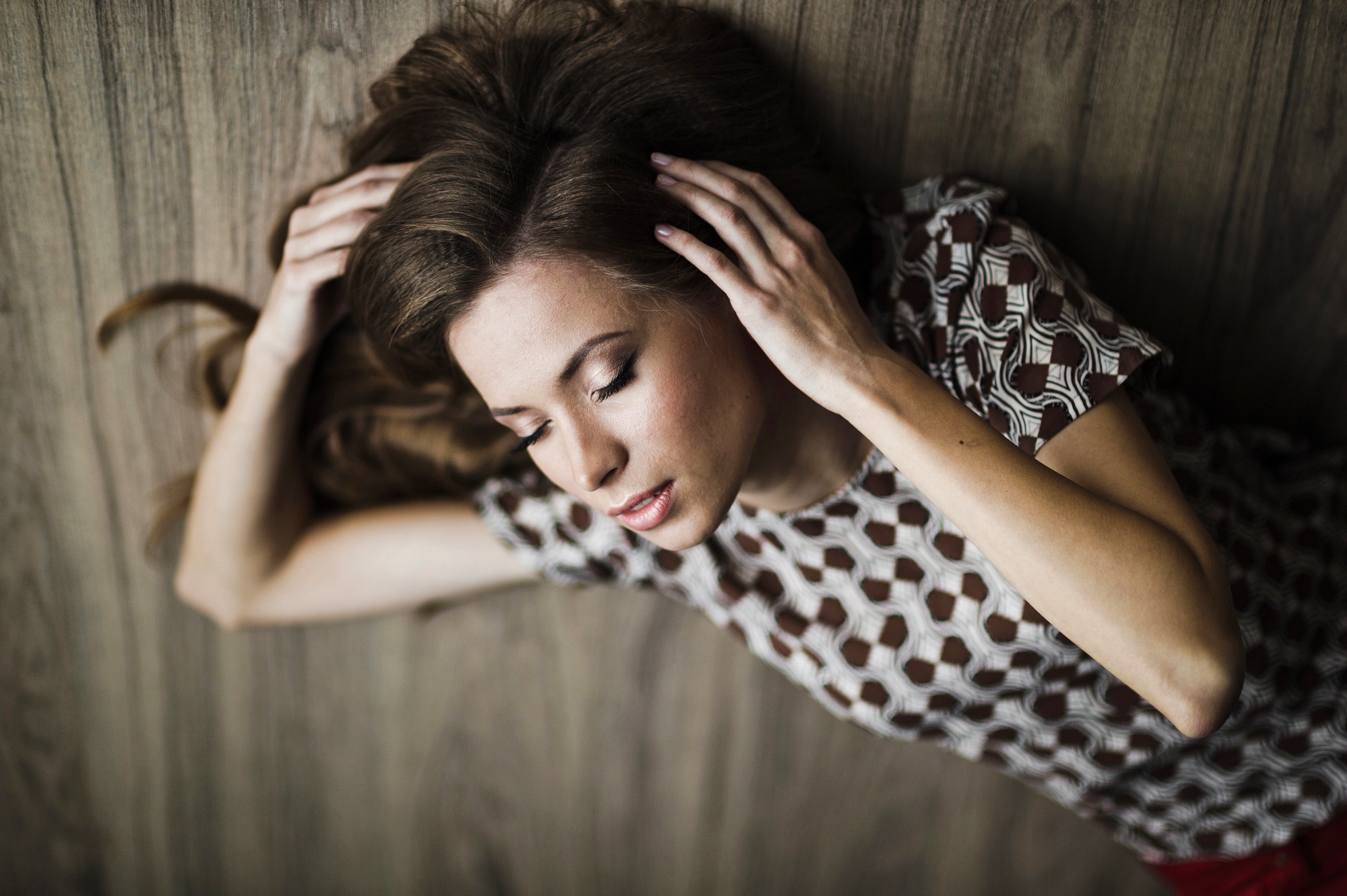 A woman with long brown hair is lying on the wooden floor with her eyes closed. She is wearing a patterned top with circular designs and has her hands gently placed on the sides of her head. The background is also wooden.