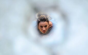 A woman with her hair tied up looks up through an abstract, circular opening. The surrounding area is blurred, drawing focus to her face and expression. She wears a blue-gray top, highlighting her inquisitive or focused expression.