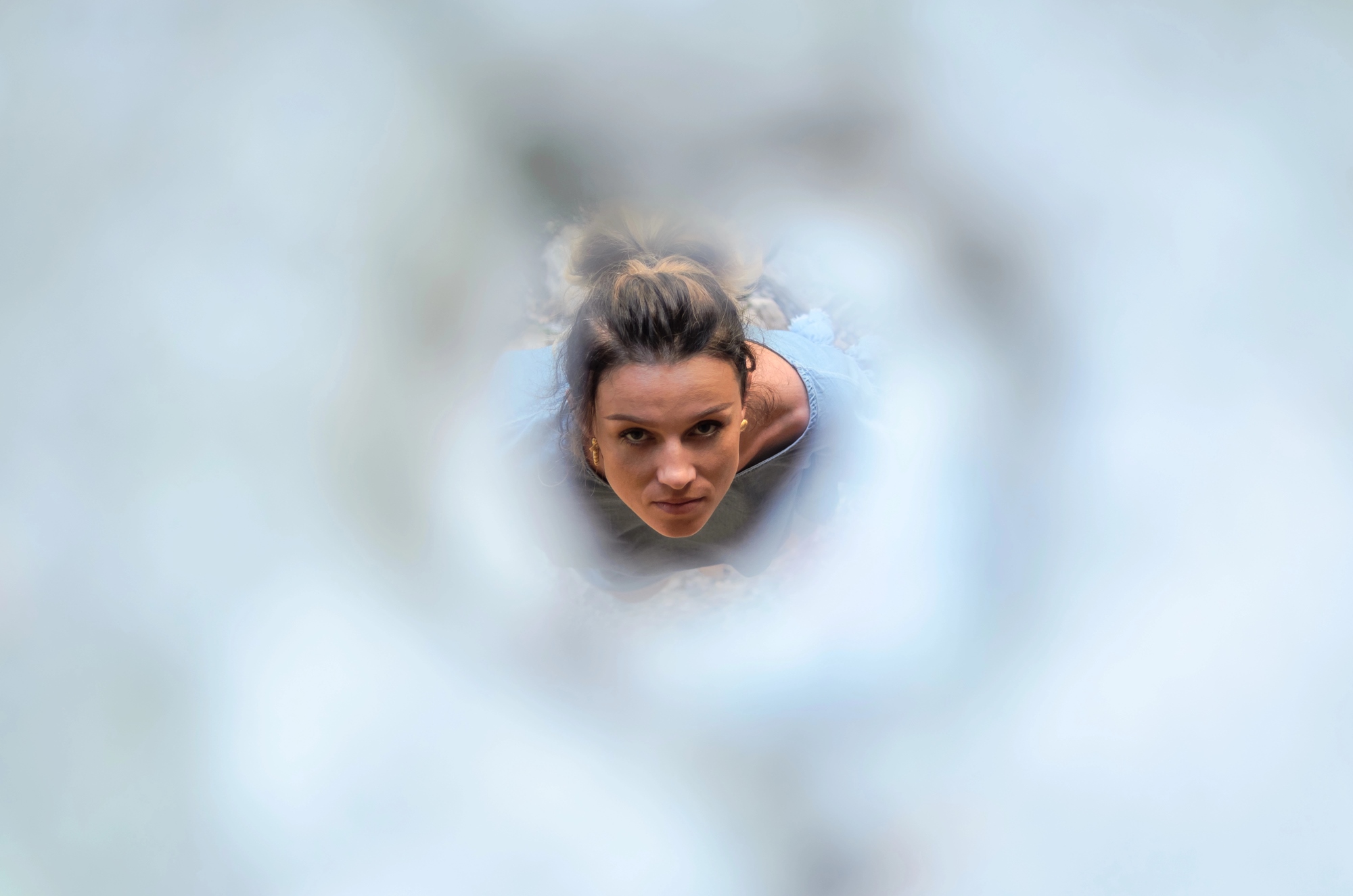 A woman with her hair tied up looks up through an abstract, circular opening. The surrounding area is blurred, drawing focus to her face and expression. She wears a blue-gray top, highlighting her inquisitive or focused expression.